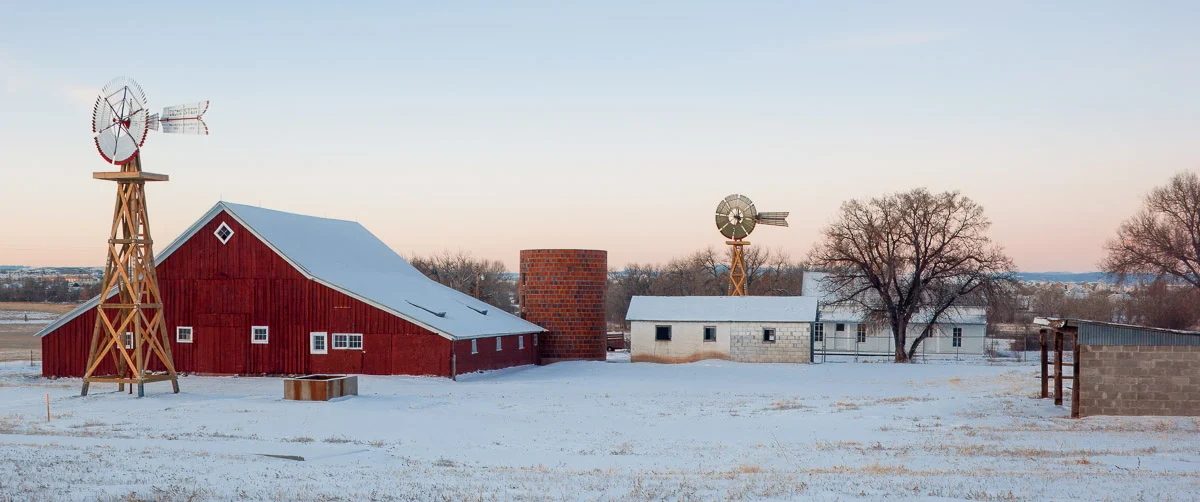 Snow-covered red barn, white farmhouse and windmill at 17 Mile House Farm Park in Colorado.